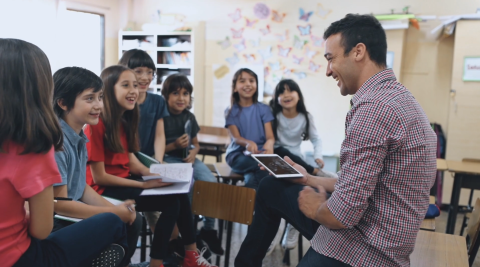 Man with iPad sits on a desk in front of a group of students.