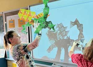 Two young white female students playing with their HyperTiles toy and casting a shadow animation on a whiteboard.